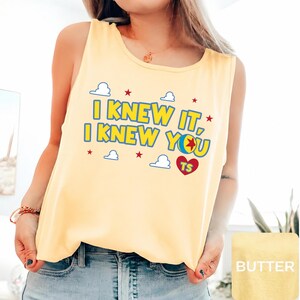 May include: A pale yellow sleeveless tank top featuring the blue text I KNEW IT, I KNEW YOU with a colourful ball graphic, red stars, white cloud outlines, and a red heart containing the letters TS printed on the front chest area.
