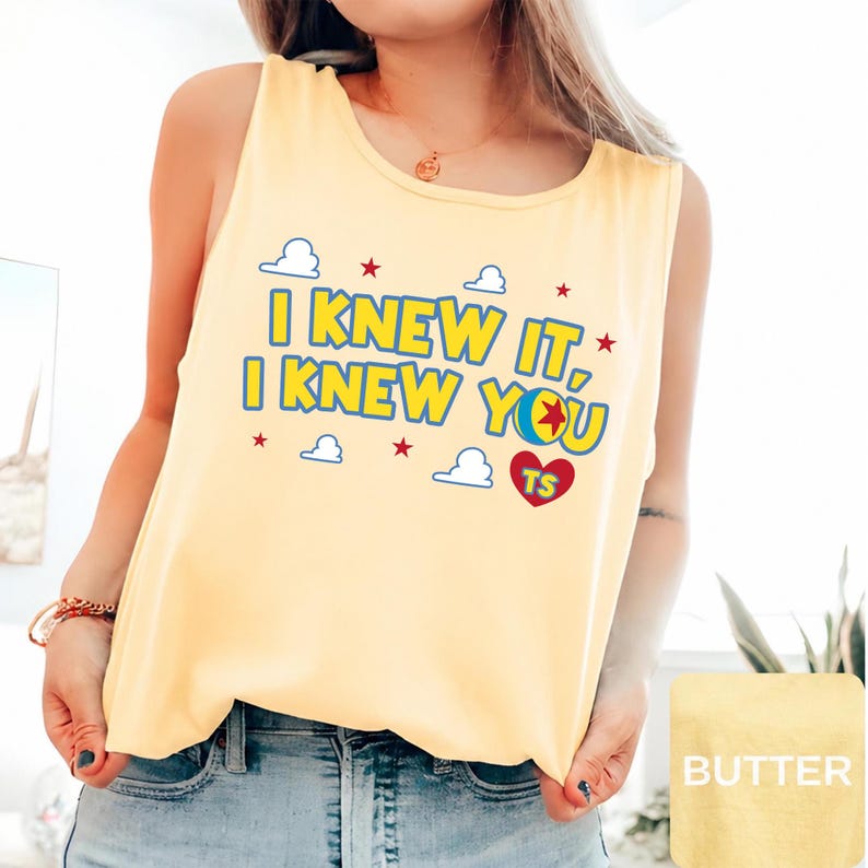 May include: A pale yellow sleeveless tank top featuring the blue text I KNEW IT, I KNEW YOU with a colourful ball graphic, red stars, white cloud outlines, and a red heart containing the letters TS printed on the front chest area.
