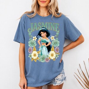 Puede incluir: Camiseta azul con un estampado de la Princesa Jasmine rodeada de flores. La palabra "JASMINE" est&aacute; impresa encima del gr&aacute;fico en verde y dorado. La camiseta es de color azul liso.