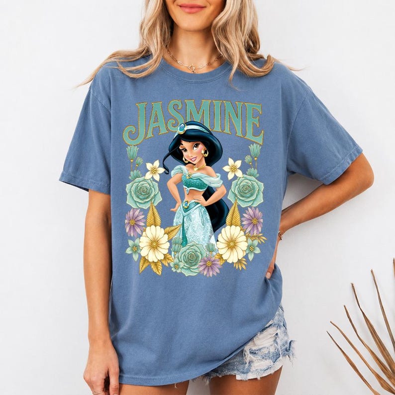 Puede incluir: Camiseta azul con un estampado de la Princesa Jasmine rodeada de flores. La palabra "JASMINE" est&aacute; impresa encima del gr&aacute;fico en verde y dorado. La camiseta es de color azul liso.