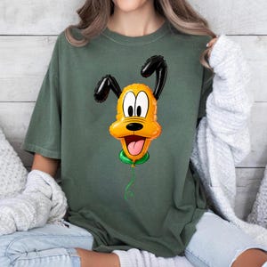 May include: A dark olive green short-sleeve t-shirt featuring a central graphic of a yellow cartoon dog face shaped like a balloon. The dog has black ears, large white eyes, a black nose, and an open mouth with a pink tongue, attached to a green string.