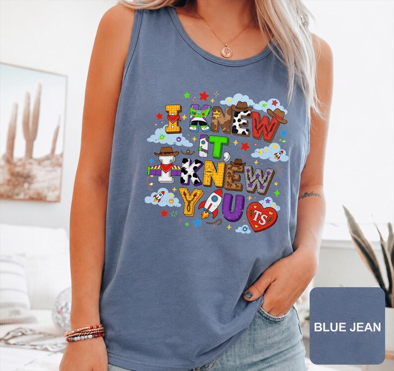 Może przedstawiać: Niebieski tank top w kolorze jeansu z kolorowym nadrukiem graficznym z napisem I KNEW IT, KNEW YOU. Wz&oacute;r zawiera r&oacute;żne motywy, takie jak łaty krowie, elementy kosmiczne i kowbojskie kapelusze, przeplatane małymi gwiazdkami, chmurami i rakietami.