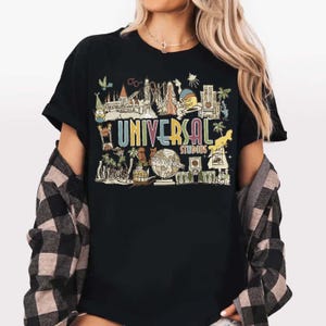 May include: Black t-shirt with a colourful graphic of Universal Studios attractions and the text "UNIVERSAL STUDIOS". The design includes cartoon illustrations of various landmarks and characters, in a vintage, retro style.