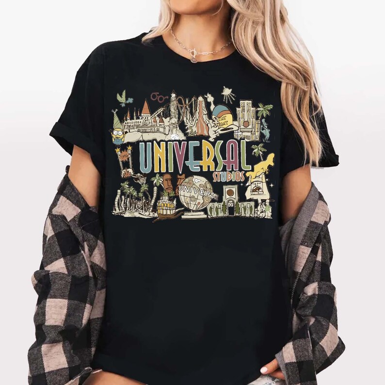 May include: Black t-shirt with a colourful graphic of Universal Studios attractions and the text "UNIVERSAL STUDIOS". The design includes cartoon illustrations of various landmarks and characters, in a vintage, retro style.