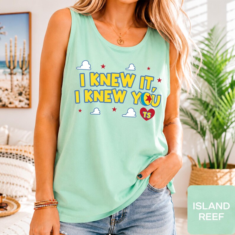 May include: A light mint green sleeveless tank top featuring the yellow text I KNEW IT, I KNEW YOU with a colourful ball graphic, small white clouds, red stars, and a red heart containing the letters TS printed on the front chest area.