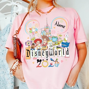 Peut inclure: T-shirt rose avec un motif Disney World color&eacute;. Le design comprend des personnages de dessins anim&eacute;s, un ch&acirc;teau et un arc-en-ciel. Le texte "Disney World 2020" est affich&eacute;. Le mot "Name" est &eacute;crit au-dessus du ch&acirc;teau.