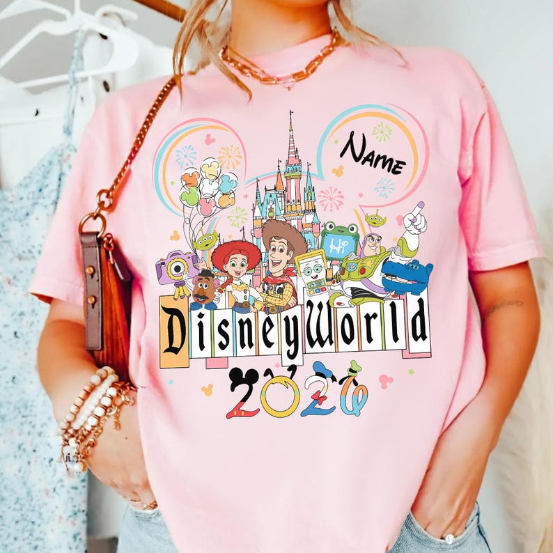 Peut inclure: T-shirt rose avec un motif Disney World color&eacute;. Le design comprend des personnages de dessins anim&eacute;s, un ch&acirc;teau et un arc-en-ciel. Le texte "Disney World 2020" est affich&eacute;. Le mot "Name" est &eacute;crit au-dessus du ch&acirc;teau.
