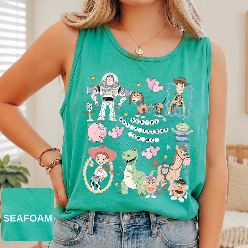 May include: A seafoam green sleeveless tank top featuring a graphic print of various Toy Story characters including Buzz Lightyear, Woody, Jessie, Rex, Slinky Dog, and others, with the text "YOU'VE GOT A FRIEND IN ME" arranged in a circular pattern.