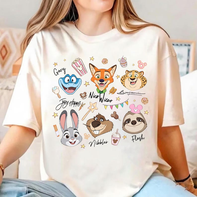 Pu&ograve; includere: T-shirt color crema con illustrazioni di personaggi dei cartoni animati. Il design include volti di animali, come una volpe, un coniglio e un bradipo, insieme a grafiche di cibo e stelle. Il testo include nomi di personaggi e frasi come "Nice Wilde".