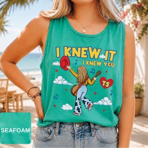 May include: A seafoam green sleeveless tank top featuring a graphic illustration of a person with long hair wearing a yellow plaid shirt and cow-print bell-bottom jeans, holding a red hat and guitar, with the text I KNEW IT I KNEW YOU and TS.