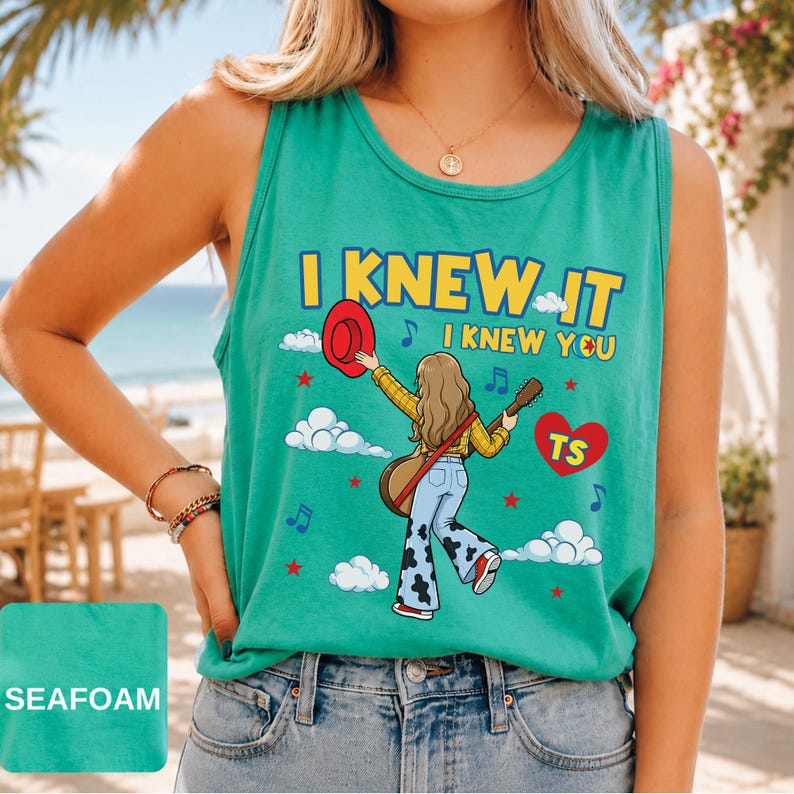 May include: A seafoam green sleeveless tank top featuring a graphic illustration of a person with long hair wearing a yellow plaid shirt and cow-print bell-bottom jeans, holding a red hat and guitar, with the text I KNEW IT I KNEW YOU and TS.