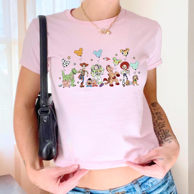 K&ouml;nnte beinhalten: Ein hellrosa T-Shirt mit kurzen &Auml;rmeln und einem Grafikdruck verschiedener animierter Figuren aus der Toy Story-Reihe, darunter Woody, Buzz Lightyear und Jessie, die jeweils einen bunten, gemusterten Ballon in Form von Mausohren halten.