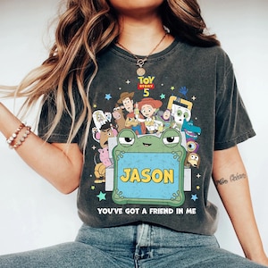 May include: Dark gray t-shirt featuring a colorful graphic of characters from Toy Story 5. A frog holds a sign that says "JASON". The text "YOU'VE GOT A FRIEND IN ME" is below the frog. The shirt is a comfortable, casual style.