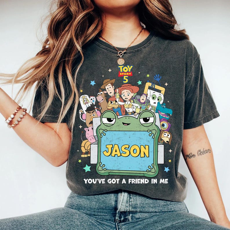 May include: Dark gray t-shirt featuring a colorful graphic of characters from Toy Story 5. A frog holds a sign that says "JASON". The text "YOU'VE GOT A FRIEND IN ME" is below the frog. The shirt is a comfortable, casual style.