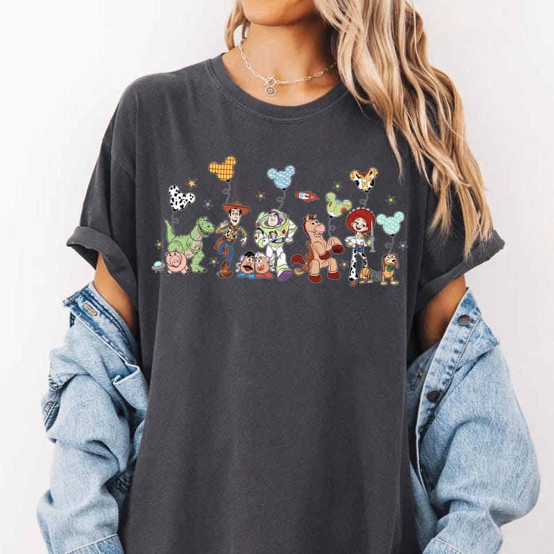 K&ouml;nnte beinhalten: Ein anthrazitfarbenes Kurzarm-T-Shirt mit einem Grafikdruck verschiedener Zeichentrickfiguren, darunter ein Cowboy, ein Weltraum-Ranger, ein Cowgirl, ein Dinosaurier, ein Schwein und ein Hund, die jeweils einen bunten Ballon in Mausohrform halten.