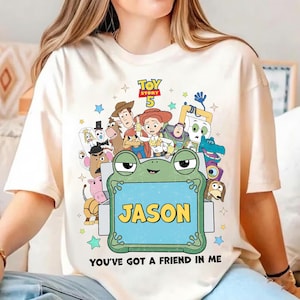 May include: Cream-colored t-shirt featuring a colorful graphic of Toy Story characters. The design includes the text "TOY STORY 5" and a frog holding a sign that says "JASON". The phrase "YOU'VE GOT A FRIEND IN ME" is printed at the bottom.