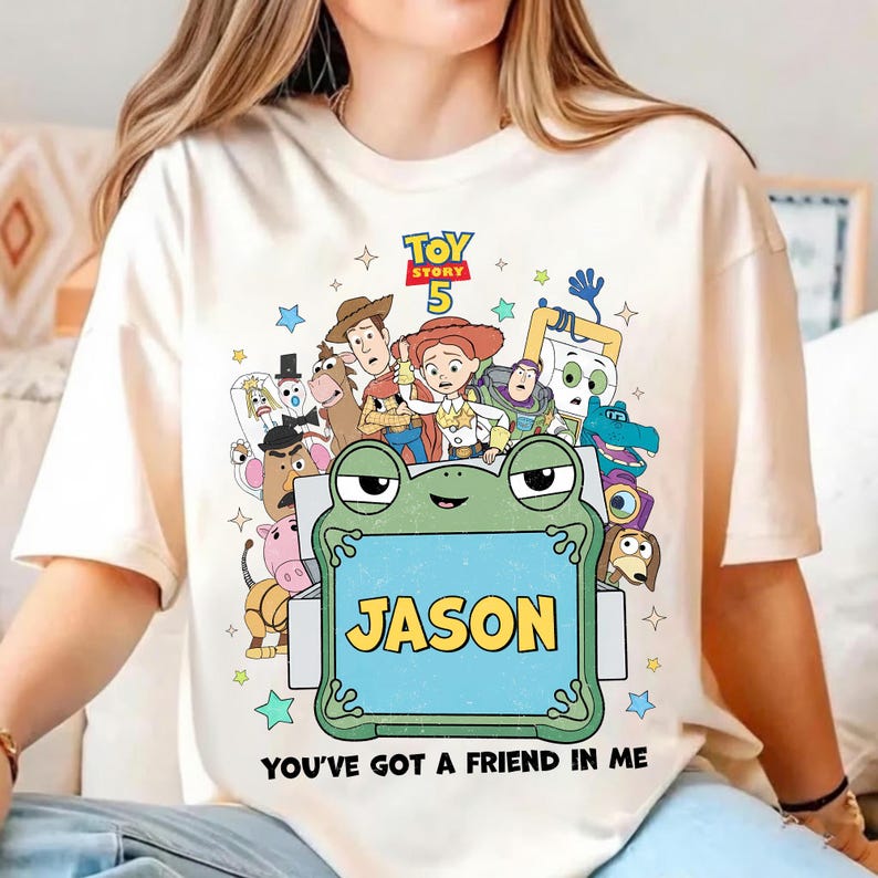 May include: Cream-colored t-shirt featuring a colorful graphic of Toy Story characters. The design includes the text "TOY STORY 5" and a frog holding a sign that says "JASON". The phrase "YOU'VE GOT A FRIEND IN ME" is printed at the bottom.