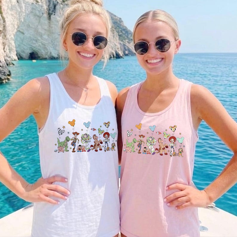 May include: Two women wearing tank tops with a Toy Story graphic. One tank top is white, the other is pink. The graphic features cartoon characters and balloons. The women are wearing sunglasses and are on a boat.
