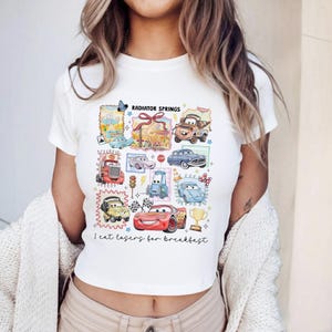 May include: White crop top featuring a colorful graphic of cartoon cars and the text "Radiator Springs" and "I eat lasers for breakfast". The design includes various car characters and elements.
