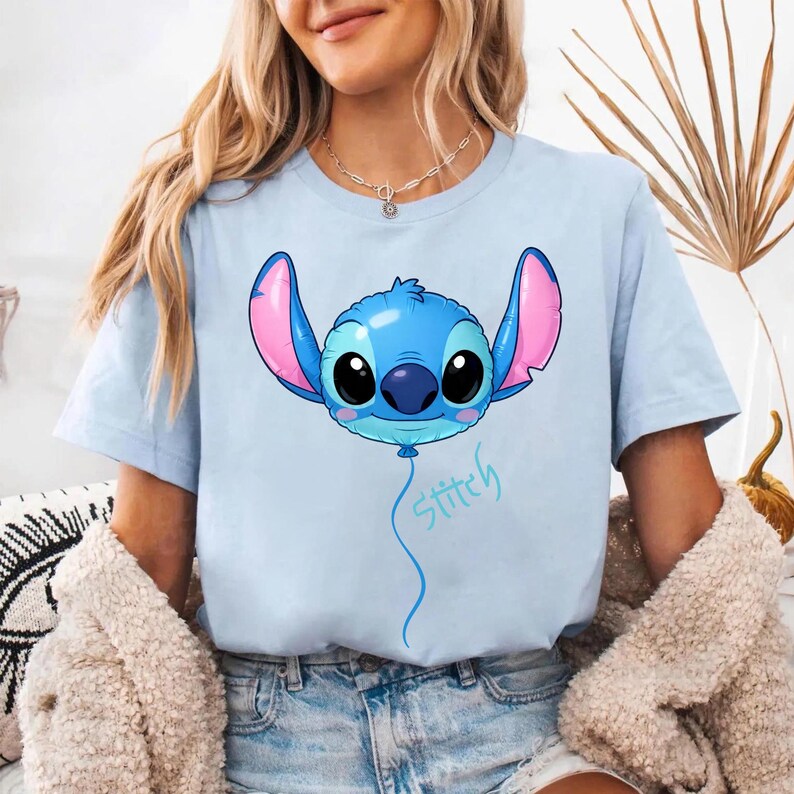May include: A light blue short-sleeve t-shirt featuring a graphic of a blue character head shaped like a balloon with large black eyes, pink inner ears, and a blue string extending downward with the word Stitch written in a light blue script font.