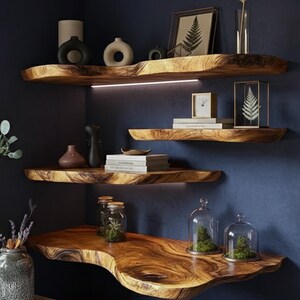 May include: Wooden floating shelves with live edge design, installed on a dark blue wall. The shelves are decorated with vases, books, framed art, and glass cloches. The wood has a warm, natural finish, creating a rustic and modern aesthetic.