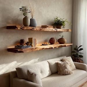 Könnte beinhalten: Zwei rustikale Holzregale mit naturbelassenen Kanten sind an einer neutralfarbenen Wand befestigt. Die Regale sind mit Vasen, Pflanzen, Büchern und Kerzen dekoriert. Unter den Regalen befindet sich ein beiges Sofa mit dekorativen Kissen.