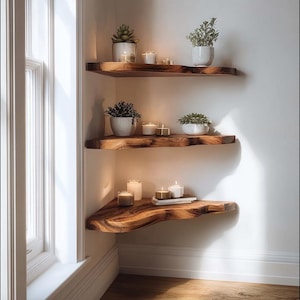 Op de afbeelding: Drie houten hoekplanken met live-edge details zijn aan een witte muur bevestigd. Elke plank bevat kleine potplanten en brandende kaarsen, waardoor een warme, uitnodigende sfeer ontstaat. De planken worden verlicht door natuurlijk licht van een raam.