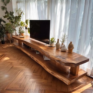 Op de afbeelding: Een grote tv-standaard van natuurlijk hout met een live edge design. De standaard heeft een onderste plank en wordt ondersteund door dikke houten poten. Er zijn verschillende decoratieve vazen en planten op geplaatst. De standaard staat tegen een achtergrond van transparante witte gordijnen.