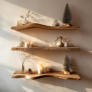 Puede incluir: Tres estantes flotantes de madera con objetos decorativos. Los estantes son de madera clara con borde natural. Los artículos incluyen velas, pequeños árboles decorativos, jarrones y un tarro de cristal con una bola decorativa.