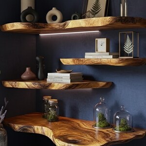 May include: Wooden floating shelves with live edge design, showcasing a natural wood grain. The shelves are adorned with decorative vases, books, and glass cloches with plants. A modern interior design element.