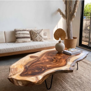 Puede incluir: Una mesa de centro de borde vivo con acabado de vetas de madera natural. La mesa está sostenida por patas de horquilla de metal negro. Rodeada por un sofá beige con cojines decorativos, un jarrón y una cesta tejida con una planta.