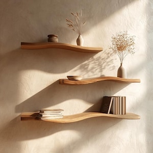 Puede incluir: Tres estantes flotantes de madera con un diseño ondulado están montados en una pared de color claro. Los estantes contienen objetos decorativos, incluyendo pequeños jarrones con flores secas, un cuenco pequeño y pilas de libros. Los estantes son de madera clara.