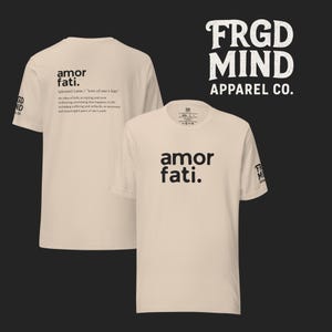 May include: Two beige t-shirts with the phrase "amor fati" printed in black. One shirt displays the definition of the phrase. The brand name "FRGD MIND APPAREL CO." is in white on a black background. The shirts are casual wear.