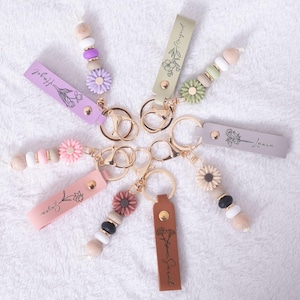 Puede incluir: Una colección de llaveros personalizados con diseños florales y nombres. Cada llavero presenta una correa de cuero con un nombre e ilustración floral, sujeta a un anillo dorado. Los llaveros tienen cuentas decorativas en varios colores, incluyendo rosa, morado y blanco.