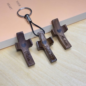 Könnte beinhalten: Drei Schlüsselanhänger mit Holzkreuzen mit eingravierten Namen und Bibelversen. Jedes Kreuz ist dunkelbraun mit einem Herz und einem Namen oder einer Schrift. Ein Schlüsselanhänger ist an einem Metallring mit einer schwarzen Kordel befestigt.
