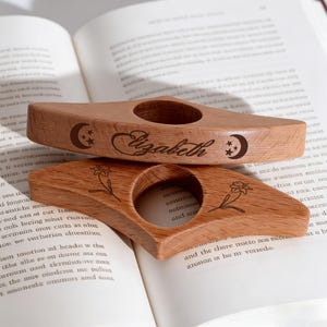 May include: Two wooden book holders with a circular opening, resting on an open book. One is engraved with the name "Elizabeth" and a crescent moon and stars. The other features a floral design. Both are made of light-coloured wood.