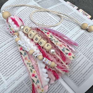May include: A handmade tassel bookmark with the name "Gabby" spelled out in wooden beads. The bookmark features pink and white fabric strips, wooden beads, and a cream-colored cord, resting on an open book.