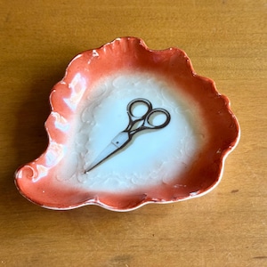 May include: A small, decorative dish with a scalloped edge, featuring a white center and a gradient orange border. A pair of small, black scissors is centered on the white surface. The dish is likely for holding small items.
