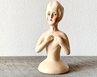 May include: A white ceramic figurine of a woman with painted blue eyes, red lips, and rosy cheeks. The figurine has her hands clasped in front of her chest and is wearing a simple white dress.