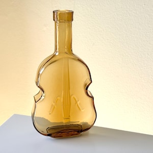 May include: A clear amber glass bottle shaped like a violin. The bottle has a narrow neck and a wide, rounded body.