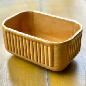 May include: A rectangular, light brown ceramic dish with vertical ribbed detailing on the sides. The dish has a smooth, glossy finish and is empty. The object is likely a butter dish or small baking dish.