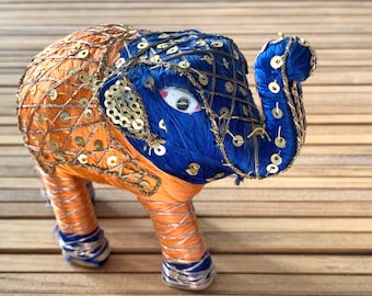 May include: A small, blue and orange elephant figurine with gold sequins. The elephant is made of fabric and has a whimsical, handmade look.