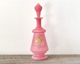 May include: A pink glass bottle with a gold floral design. The bottle has a stopper and a round base.
