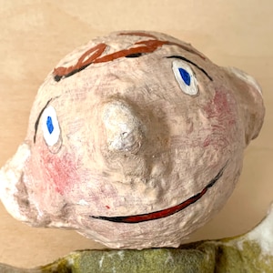 May include: A handmade papier-mâché sculpture of a smiling face. The face is painted with blue eyes, red cheeks, and a red smile. Brown paint accents the top of the head. The sculpture has a textured, off-white finish.