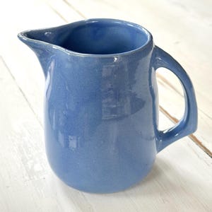 May include: A blue ceramic pitcher with a handle and a spout. The pitcher has a rounded body and a smooth, glossy finish. The pitcher is approximately 7 inches tall and 5 inches wide.