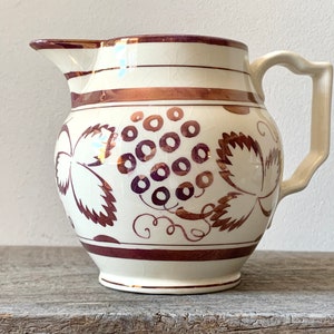 May include: Cream-colored ceramic pitcher with a brown grape design. The pitcher has a curved handle and a spout. The grape design includes clusters of grapes and leaves. Brown bands encircle the top and base of the pitcher.