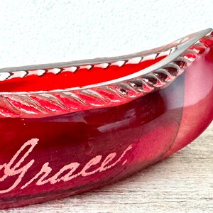 May include: A red glass canoe-shaped dish with a rope-like design around the rim. The dish is inscribed with the word "Grace".