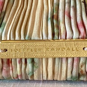 Close-up of Loeffler Randall gold logo plaque on tan floral pleated fabric clutch.