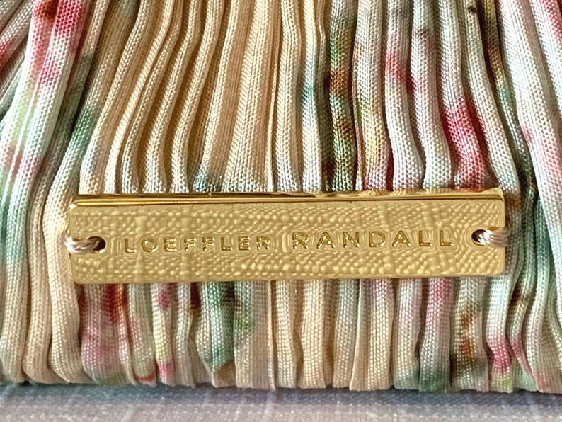 Close-up of Loeffler Randall gold logo plaque on tan floral pleated fabric clutch.