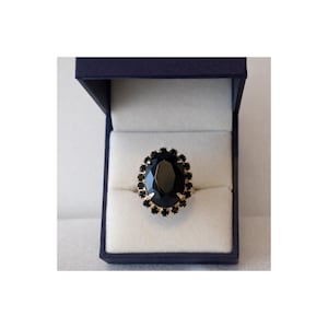 Op de afbeelding: Een goudkleurige ring met een grote, ovale zwarte edelsteen, omringd door kleinere zwarte stenen. De ring wordt gepresenteerd in een marineblauwe ringdoos met een witte binnenkant.